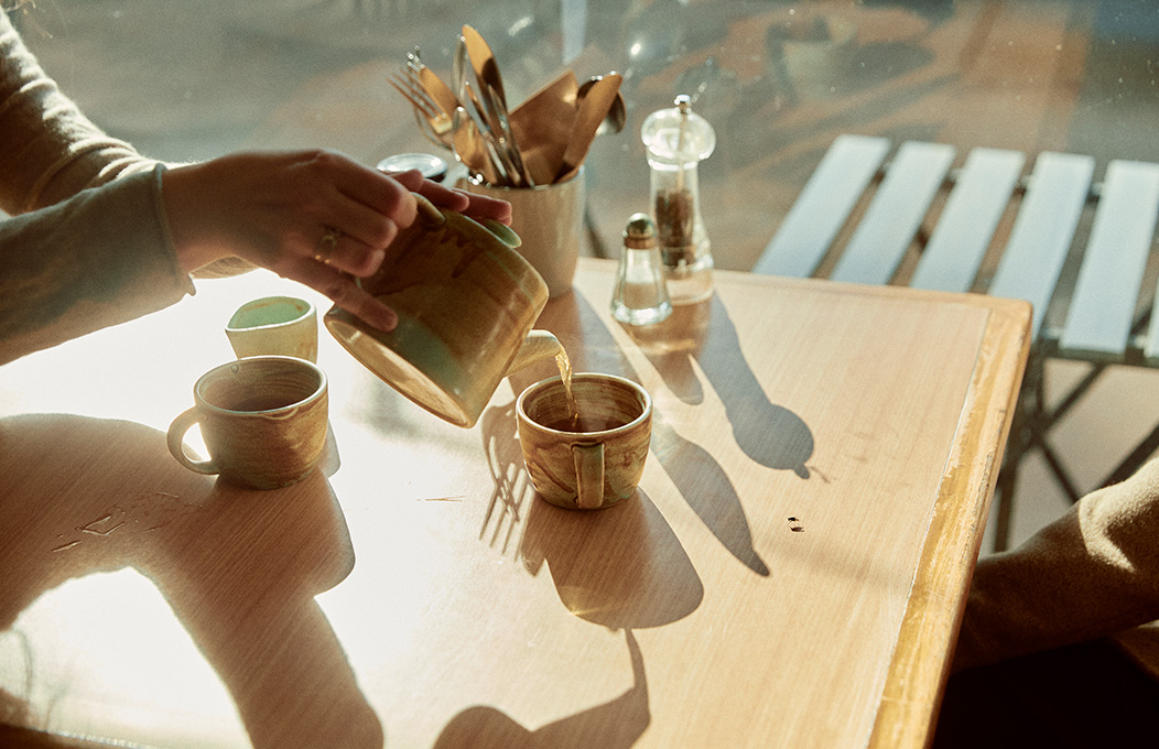 Image of customers sitting around a square table at a cafe next to the window with one pouring milk into a coffee cup on the table,