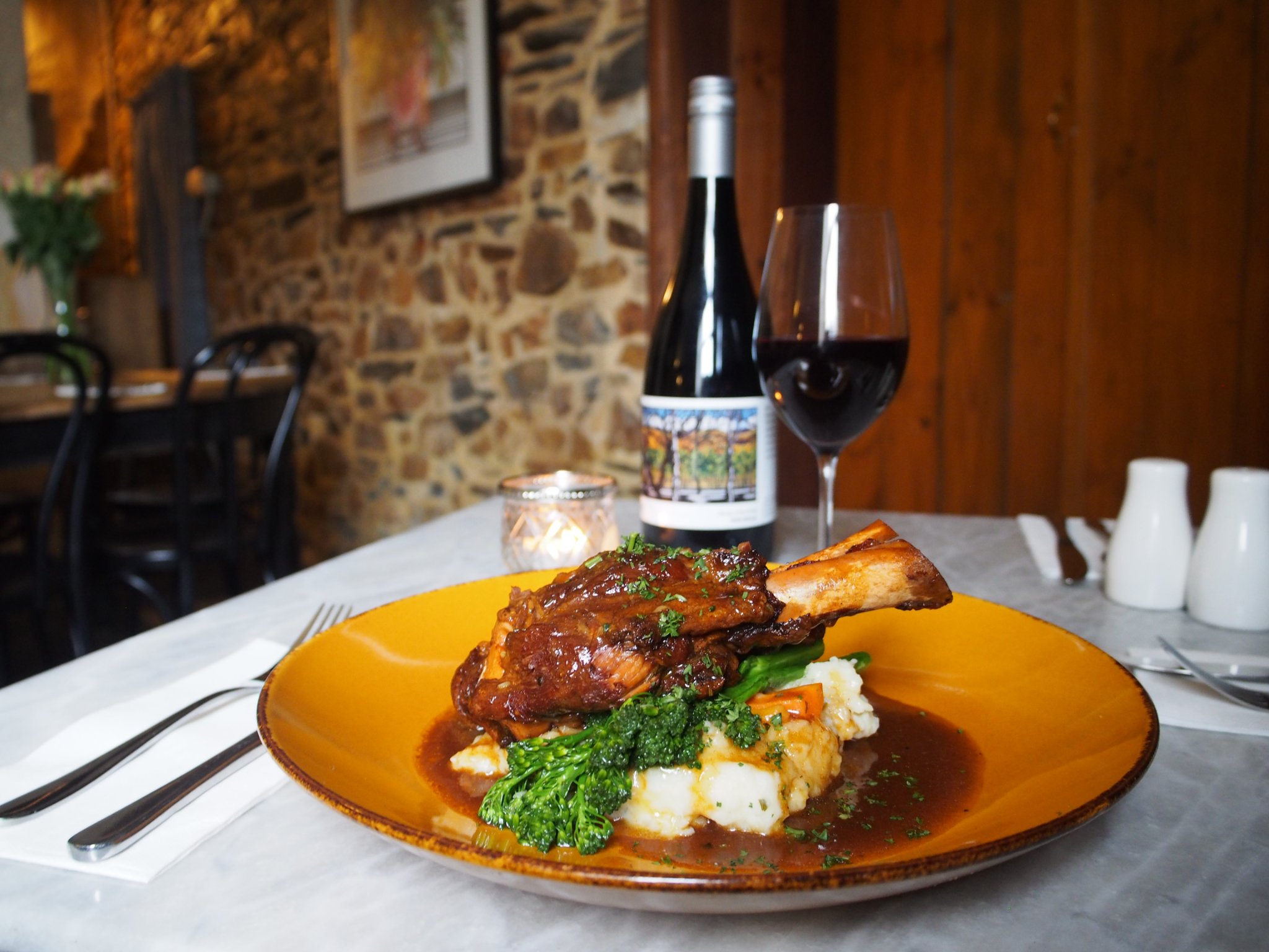 A table dressed with a plate of slow cooked lamb shank and veggies, a glass and bottle of red wine, sitting on a white table cloth.