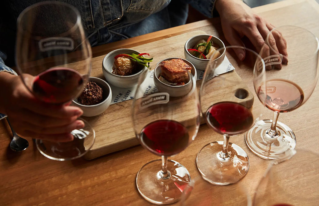 A wooden table set for wine tasting, featuring four glasses of red wine and small bowls of assorted appetizers on a serving board, with hands mid-pour.
