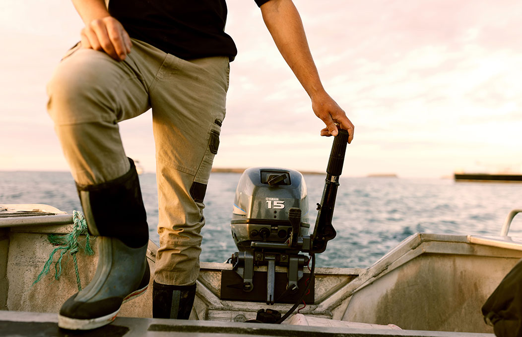 Person standing in a tinny holding the tiller of an outboard motor with calm ocean behind.