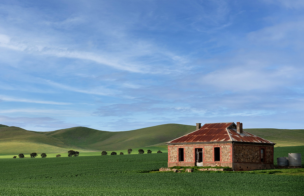 A paint image of the Burra Mines site, with green pains and a blue sky, and the brick style building on the right-hand side of the image.