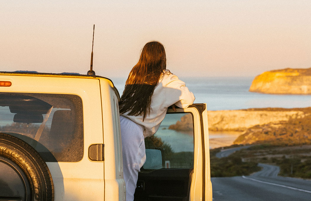 Person leaning out of a 4WD door overlooking a winding coastal road and ocean cliffs at sunset.