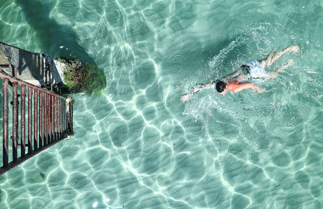 Surrounded by clear turquoise water, someone swims up to the ladder of the Jetty at Glenelg 