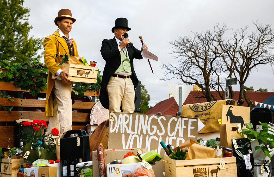 Two people in vintage attire stand on a wooden platform at an outdoor vintage market, surrounded by crates of produce and flowers, hosting a lively auction event.