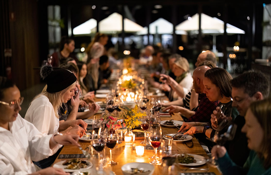 Guests sitting on each side of the table, eating and enjoying winter red vines, with lightful candles in the center of the table.