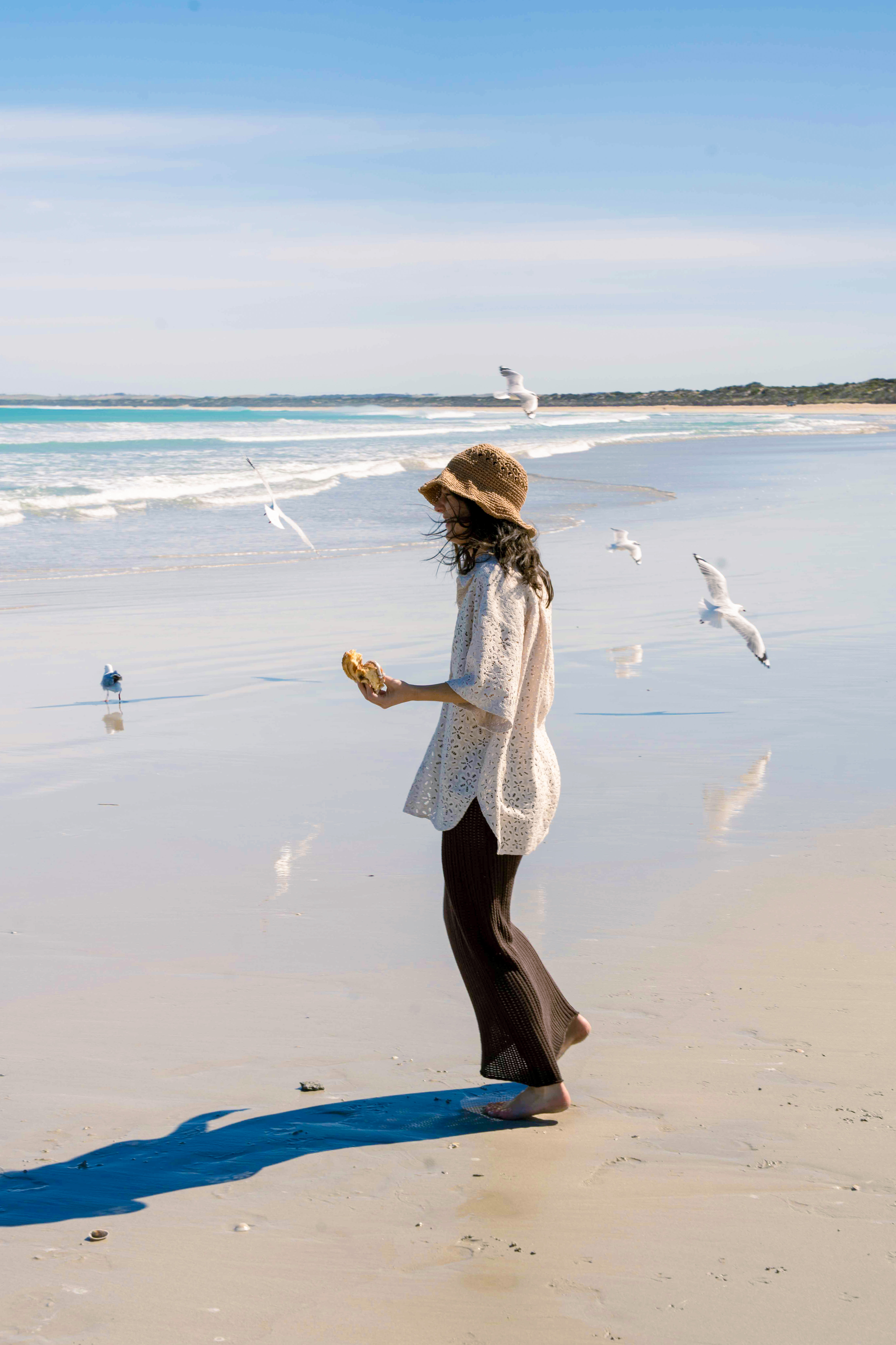woman in a sun hat laughs at the beach with a scroll in hand, seagulls flying around her in the air