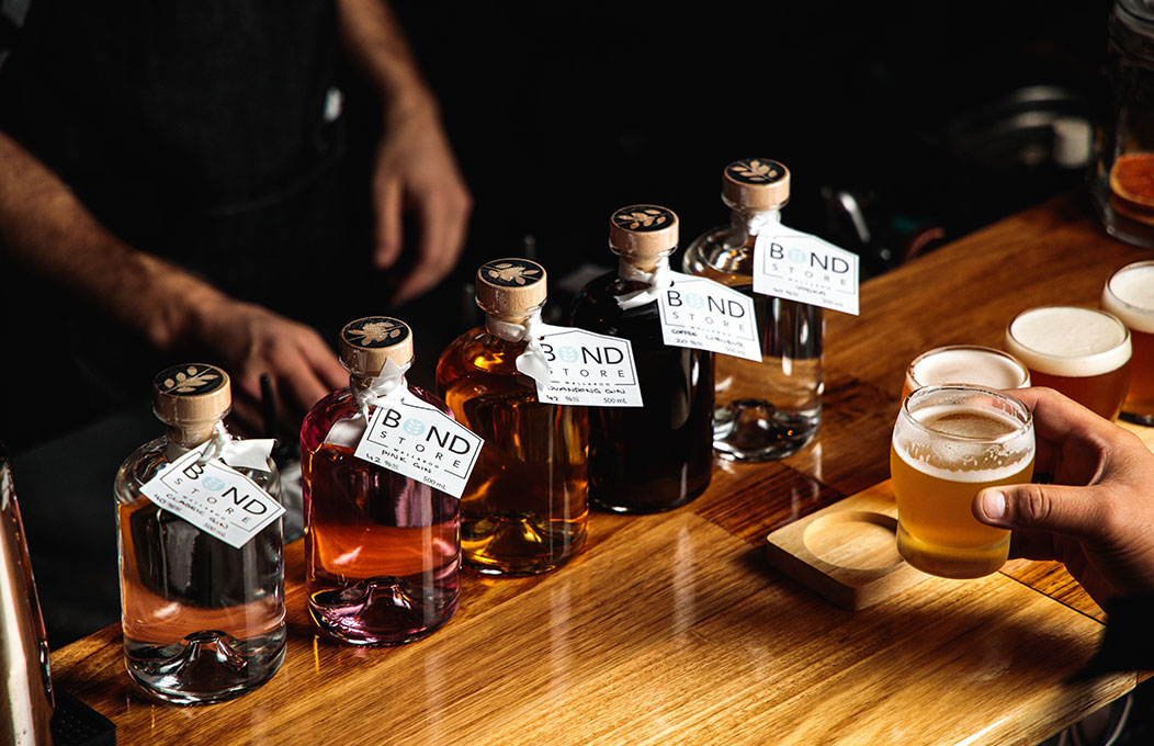 Row of Bond Store spirits on a wooden bar with beer glasses and a hand holding a drink.