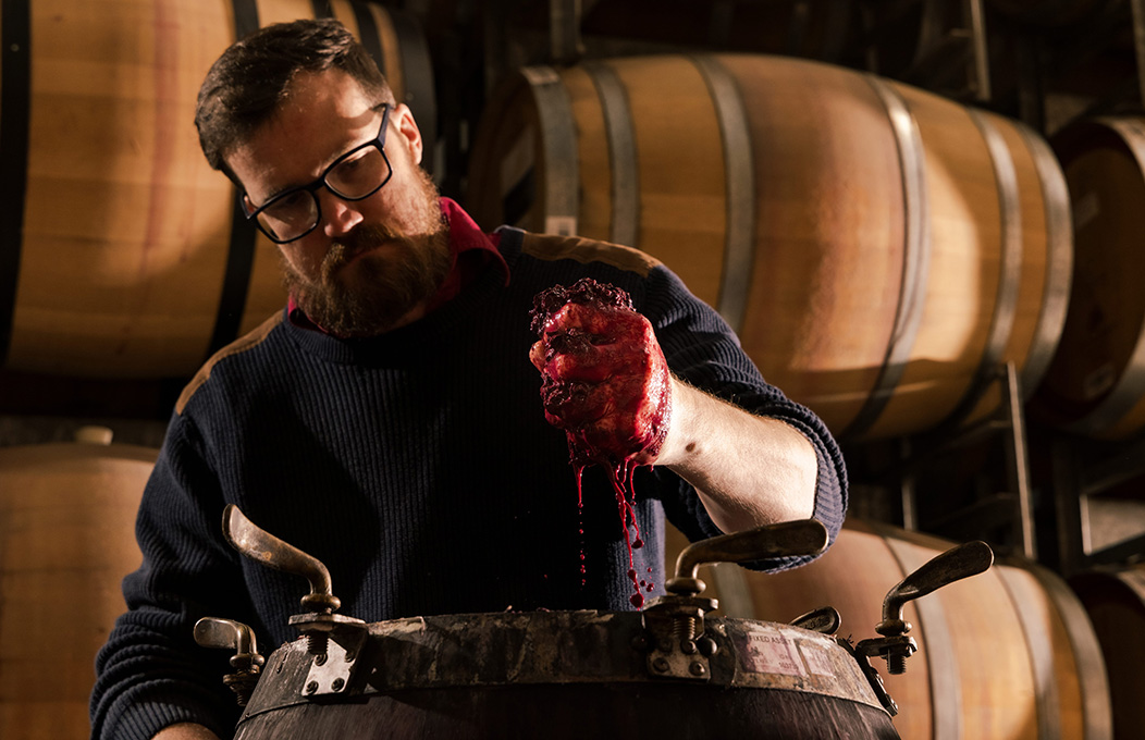 A vigneron squeezing grappe on top of an oak barrel, with other barrels decorating the background of the image.