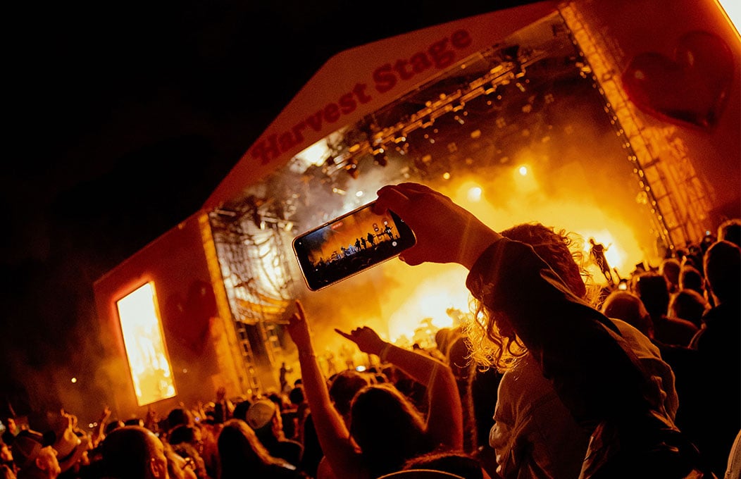 Someone holding up their phone in a crowd of music lovers captures the stage of Harvest Rock which is bathed in a yellow glow