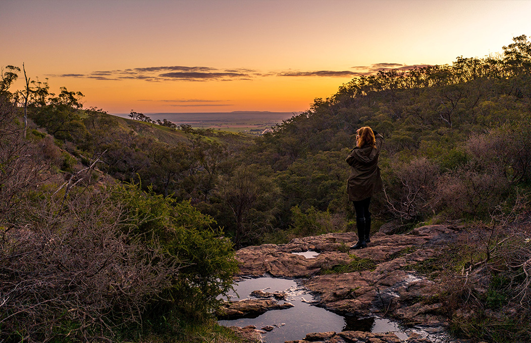 A lady looking at a sunset view from a hike setting perspective, with a water place in the foreground that seems to be the top of a waterfall.