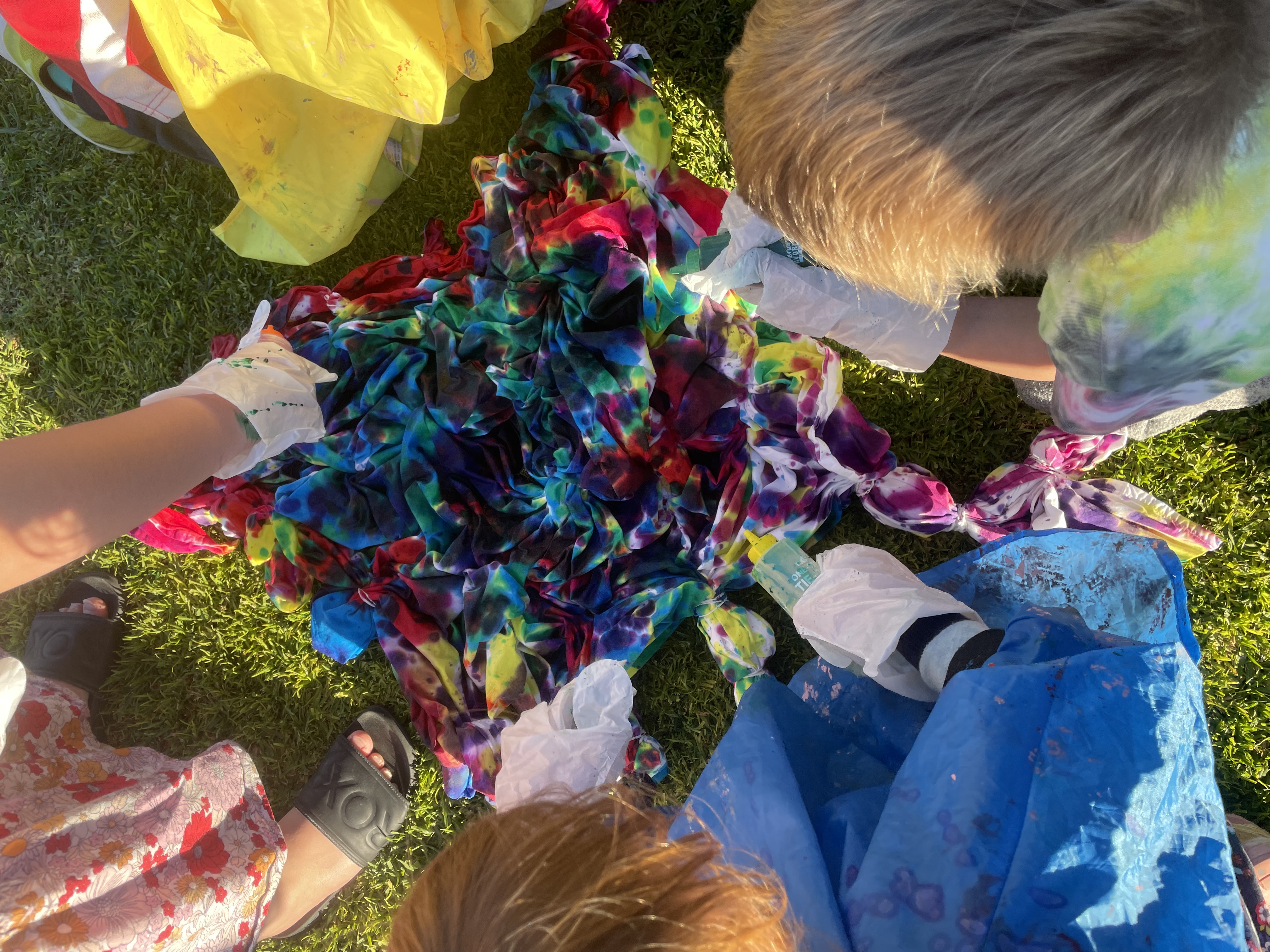 Kids outdoors on grass creating vibrant tie-dye patterns, with gloved hands working on crumpled fabric dyed in bright reds, blues, greens, and purples