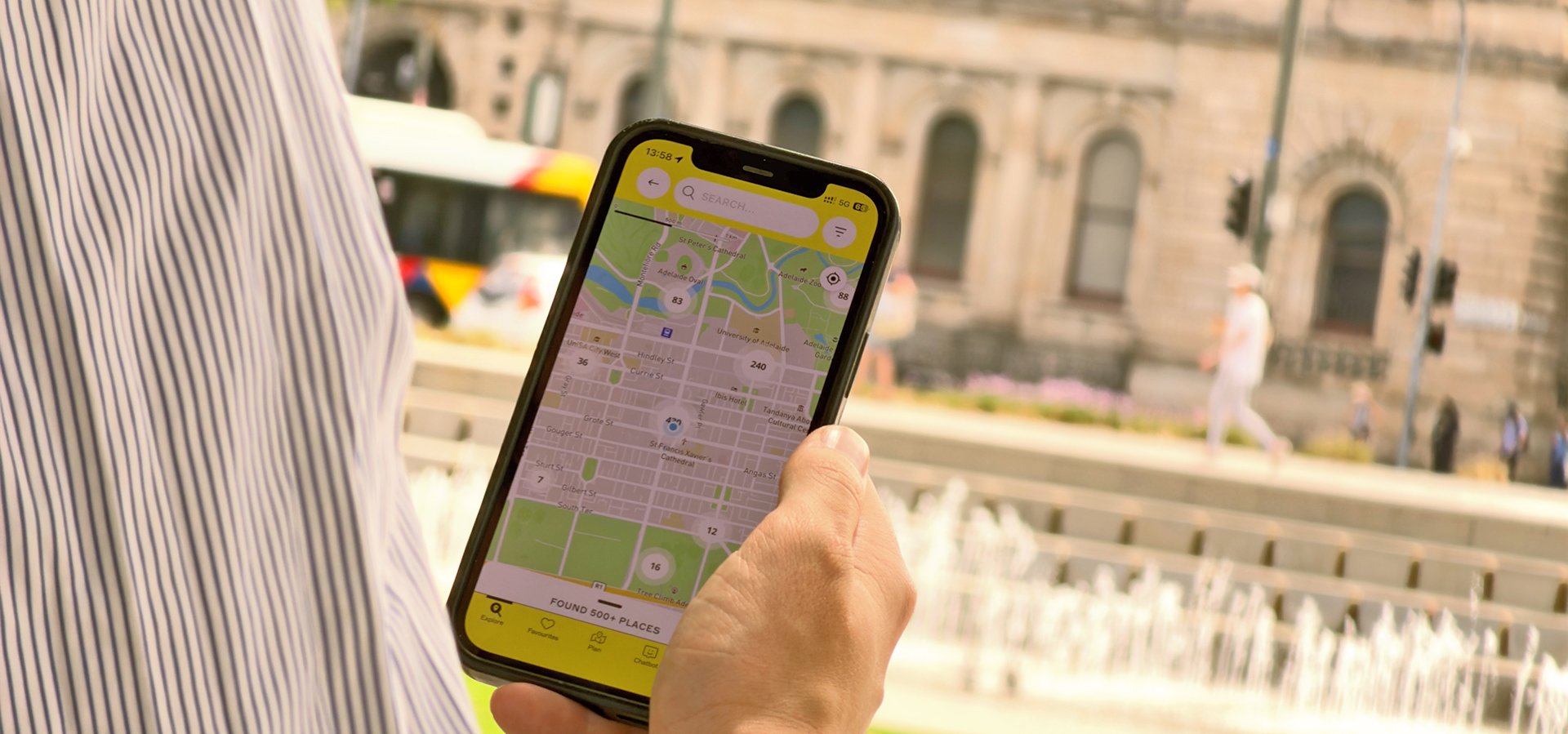 Peron holding a phone displaying the map view of the Explore South Australia app with Victoria Square water jets and Adelaide Metro bus in the background