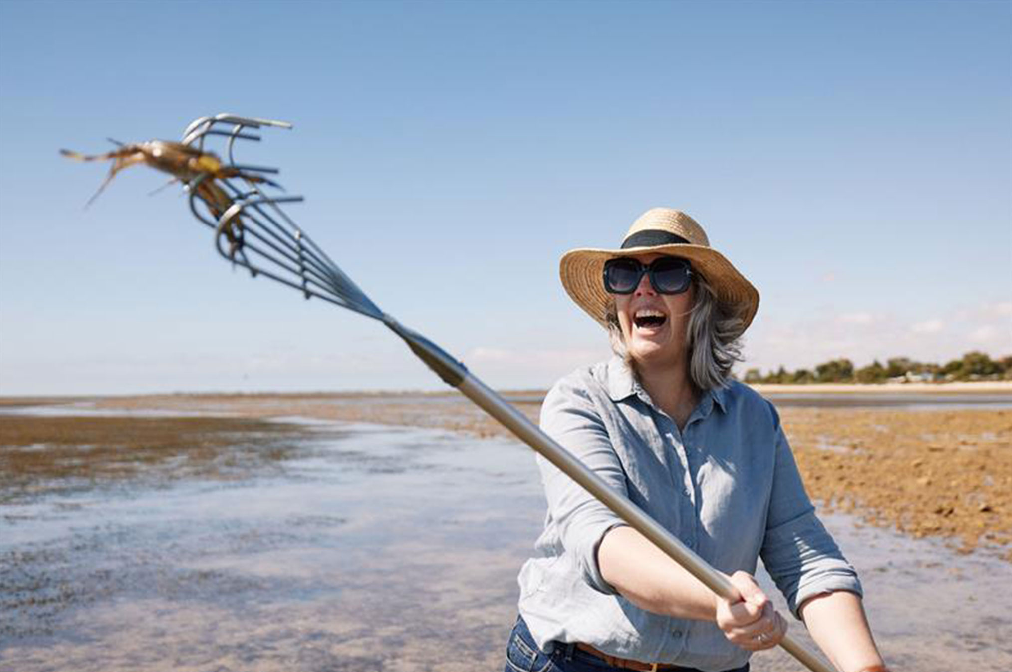 Person holding a rake with a freshly caught crab on a wide tidal flat under a bright blue sky, with shallow water and distant shoreline in view.