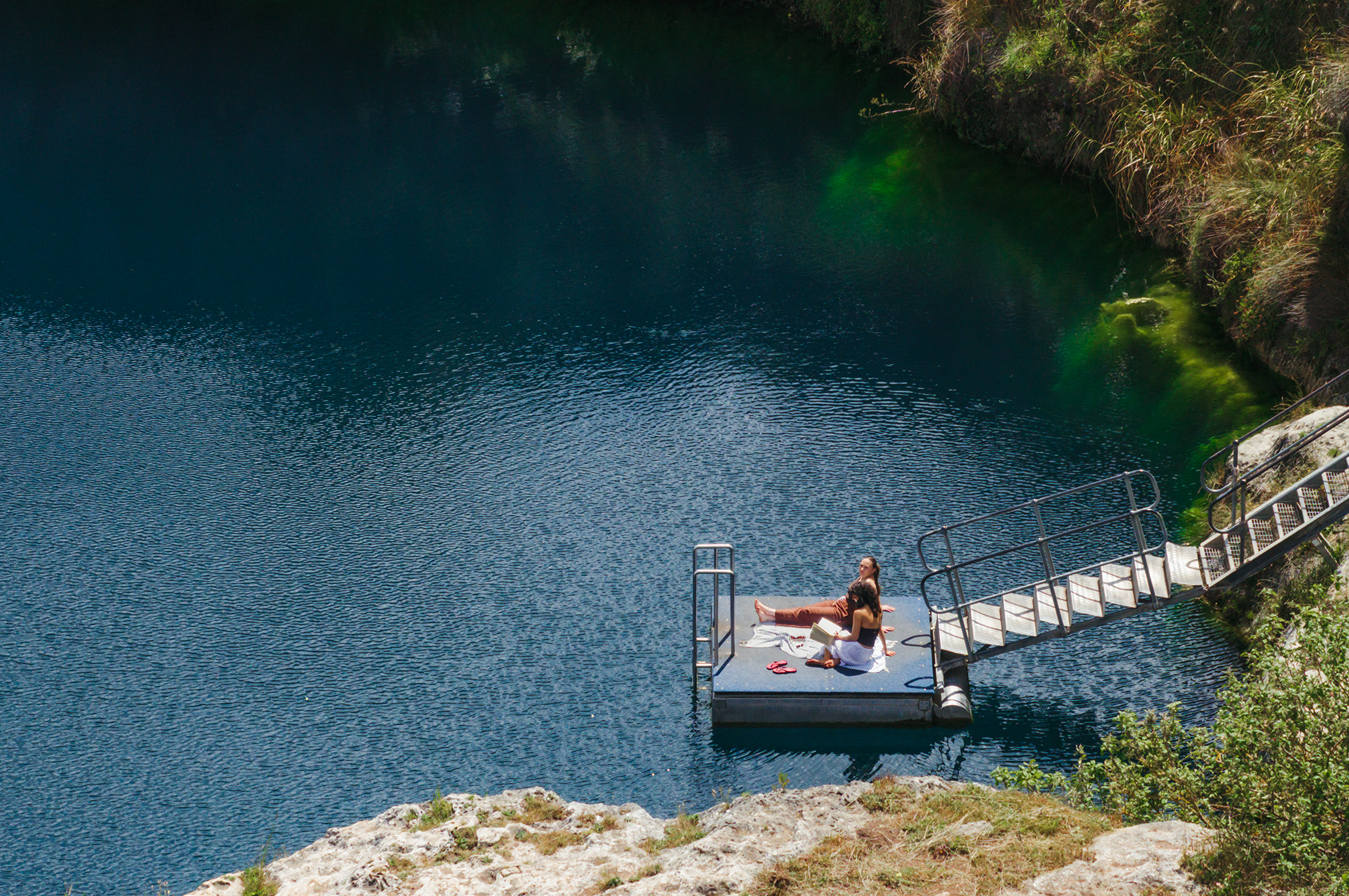 Blue water surrounds two people who relax on the pontoon at Little Blue Lake on the Limestone Coast 